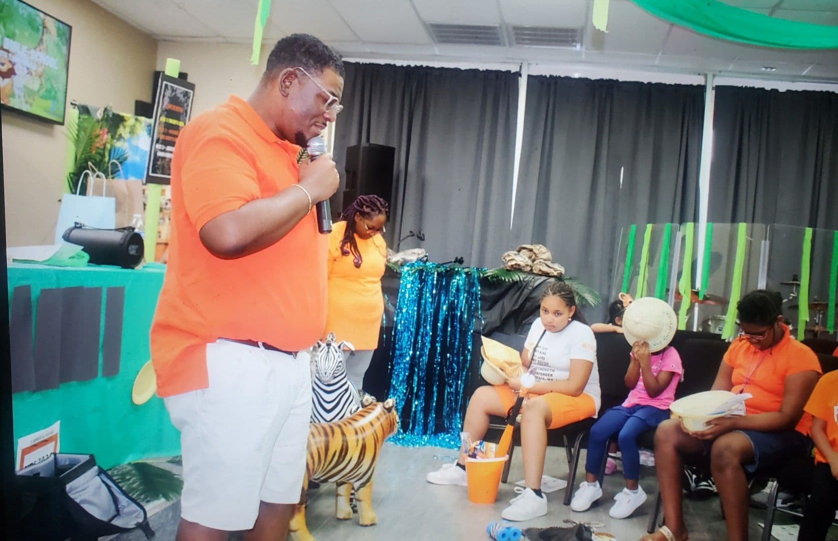 A leader in an orange shirt speaking into a microphone to kids at VBS